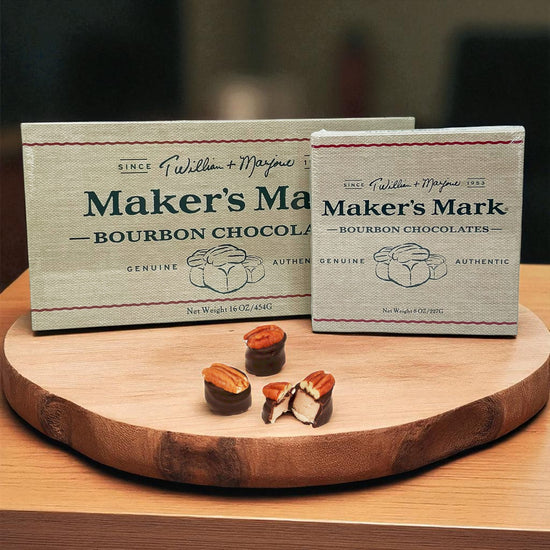 Two boxes of Maker's Mark Bourbon Chocolates on a wooden board with a blurred background.
