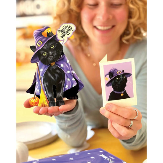 Person holding a black cat figurine in a witch costume with a 'Happy Halloween' card.