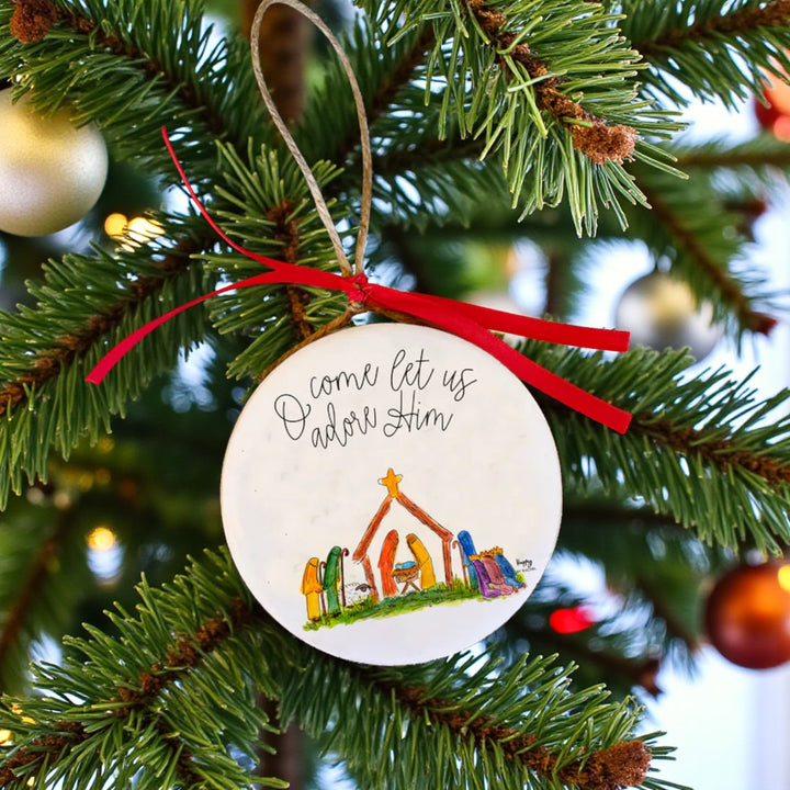 A white circular Nativity ornament with the words "O come let us adore Him" hanging on a Christmas tree.