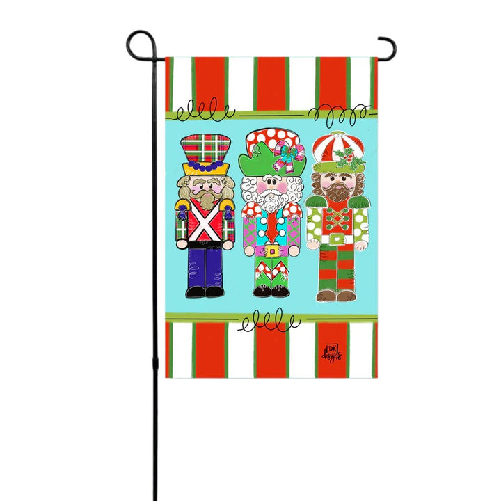 A colorful holiday garden flag with three whimsical nutcracker soldiers hanging on a black stand.
