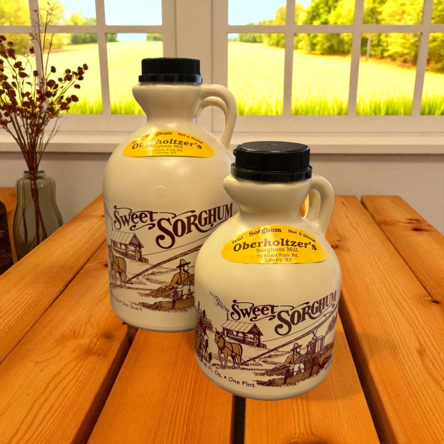 Two Oberholtzer’s Sweet Sorghum jugs, 32oz and 16oz, on a wooden table by a window with a farm scene label.