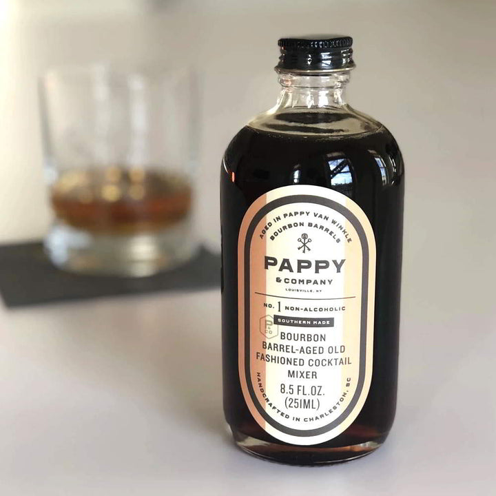 Bottle of Pappy & Company non-alcoholic bourbon-aged old fashioned cocktail mixer on a white surface with a glass in the background.
