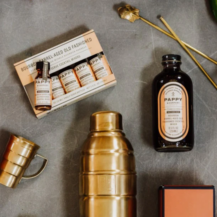 Pappyоварio bottles and packaging on a gray surface with a gold shaker and mugs.