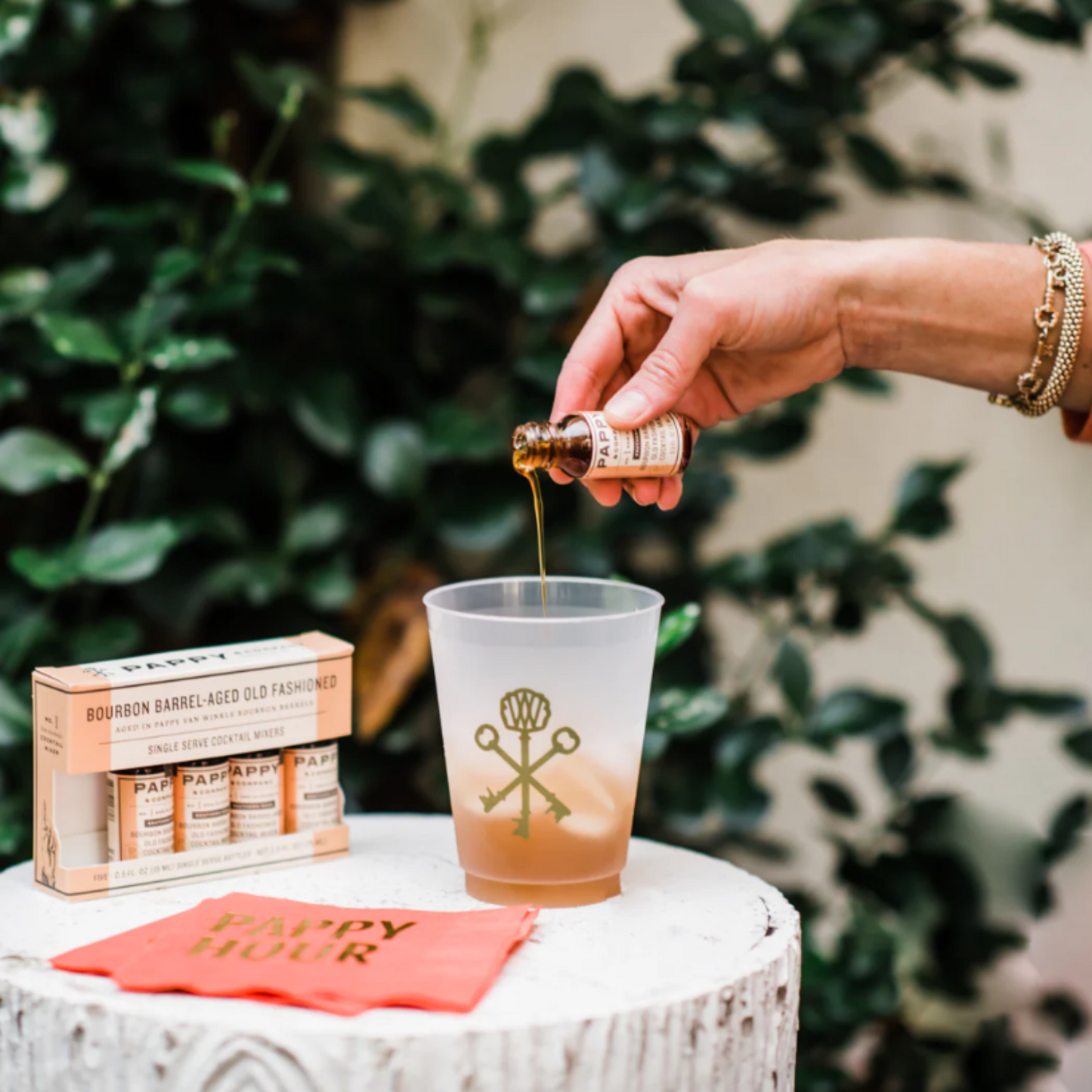Person pouring a drink into a glass with a decorative label, surrounded by a box and napkins on a table.