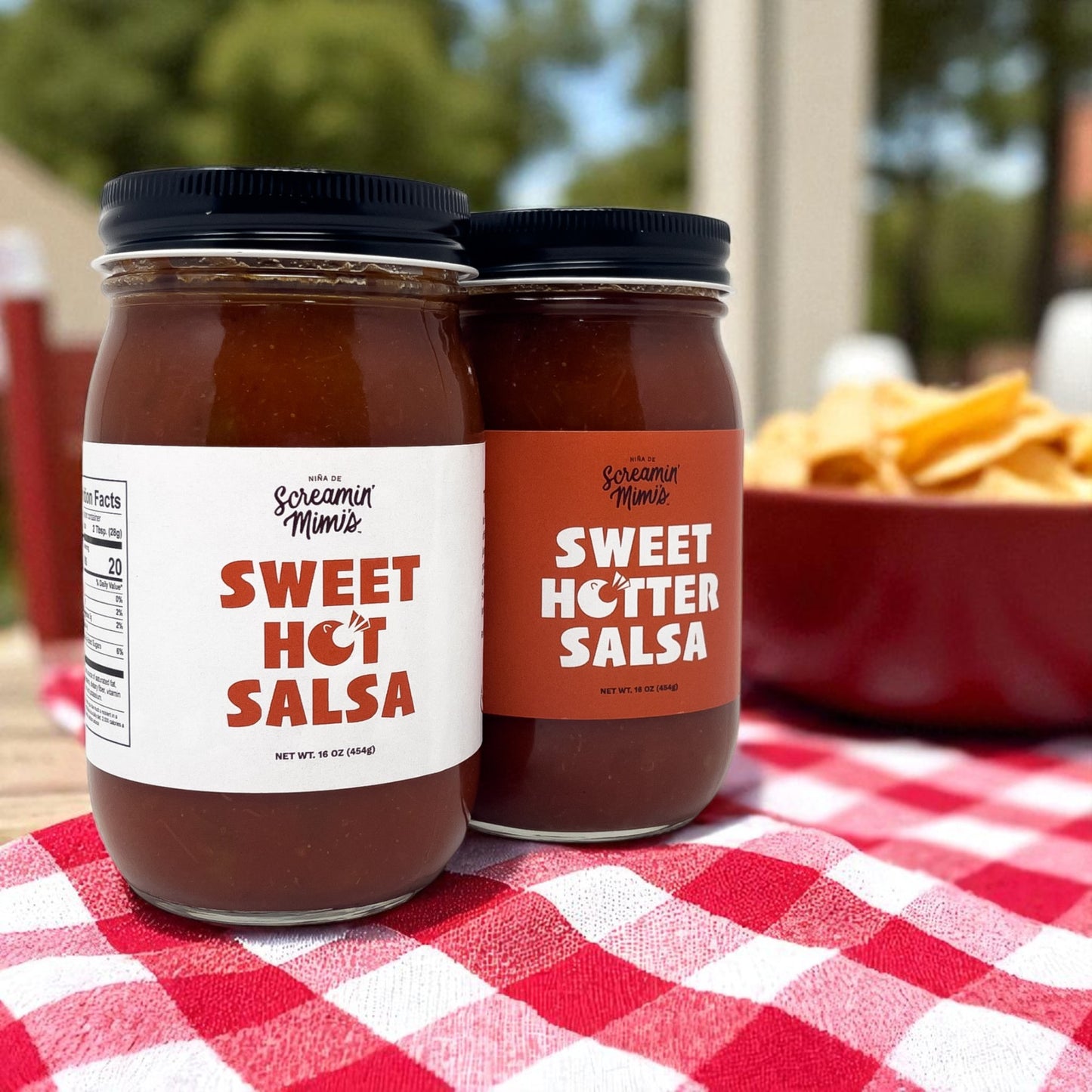 Two jars of Sweet Hot Salsa on a red and white checkered tablecloth with chips in the background.