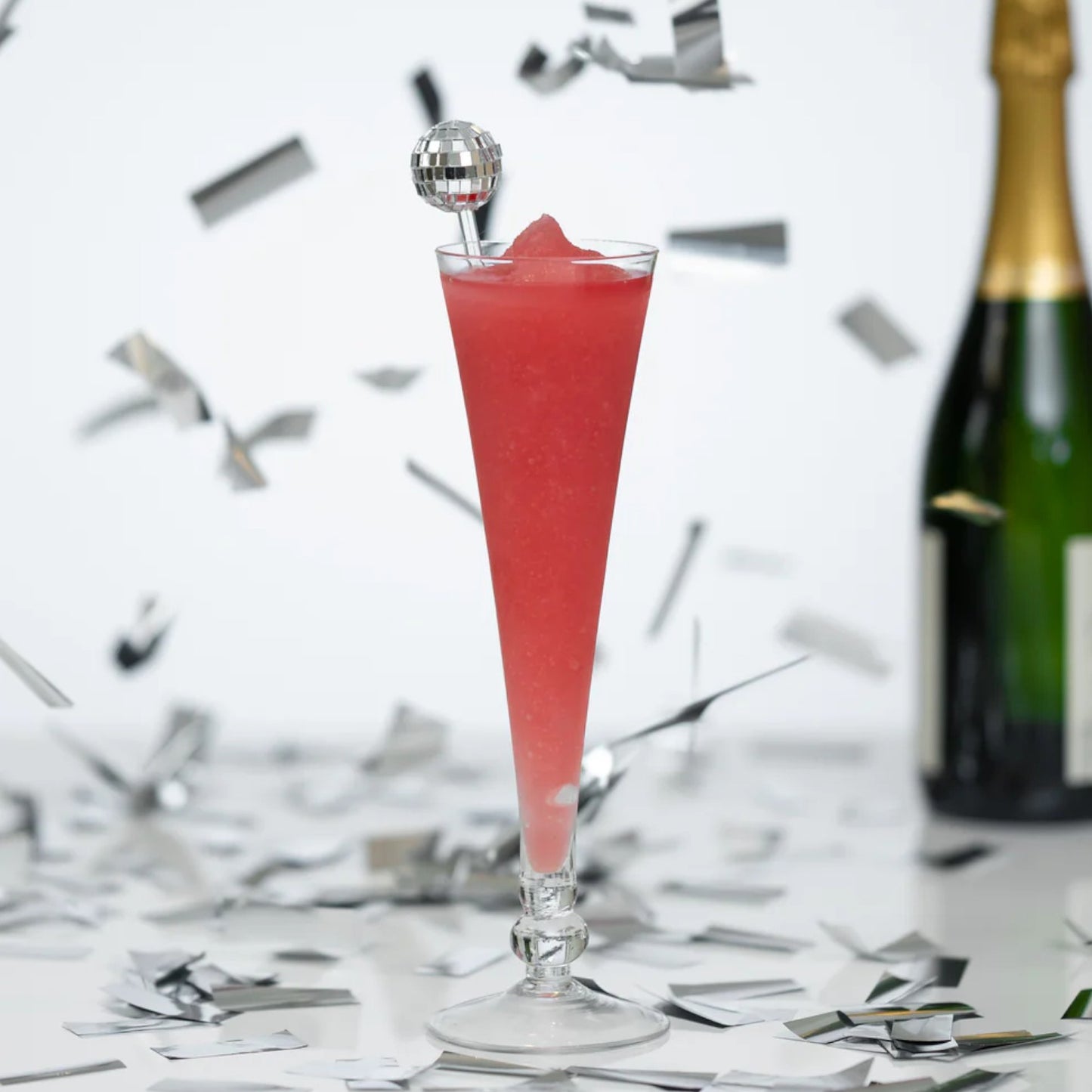 Pink cocktail in a glass with a bottle of champagne on a white background with silver confetti.