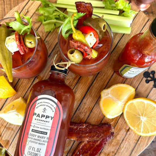 Two Bloody Mary cocktails with garnishes on a wooden board with Pappy & Co. Bloody Mary mix bottle.