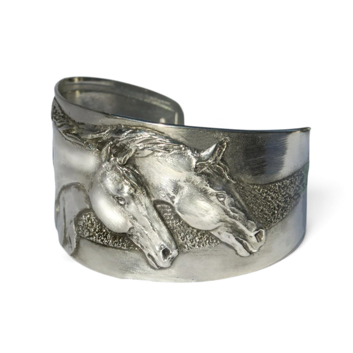 Studio shot of the Two Horse Heads Pewter Cuff Bracelet in silvery metal on a white background, showing the detailed raised relief design of two running horses.