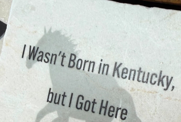 Coaster with horse silhouette and text 'I Wasn't Born in Kentucky, but I Got Here' on a wooden surface.