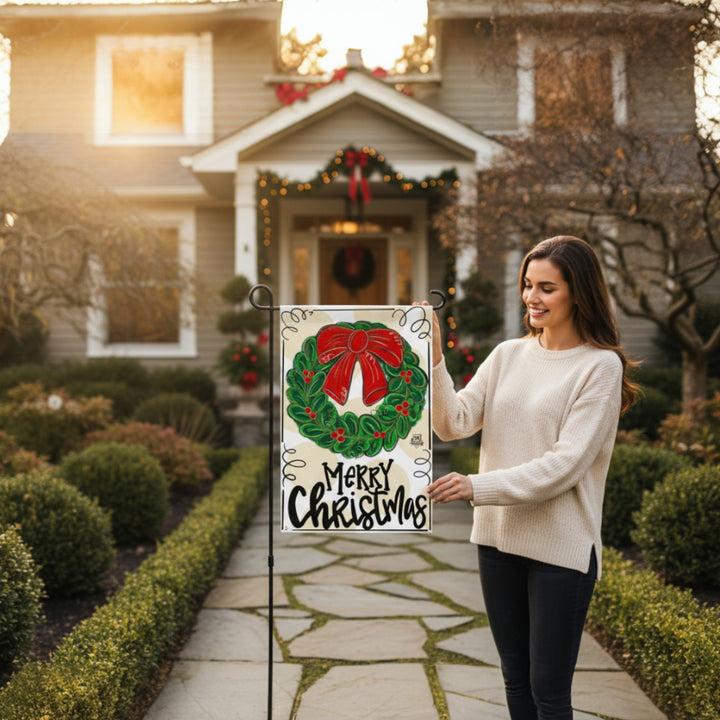 A woman smiling while holding the Merry Christmas Wreath garden flag in front of her home.