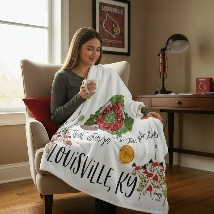 Young woman sitting comfortably in a beige armchair, sipping from a white mug, and covered by the Louisville Game Day fleece blanket.