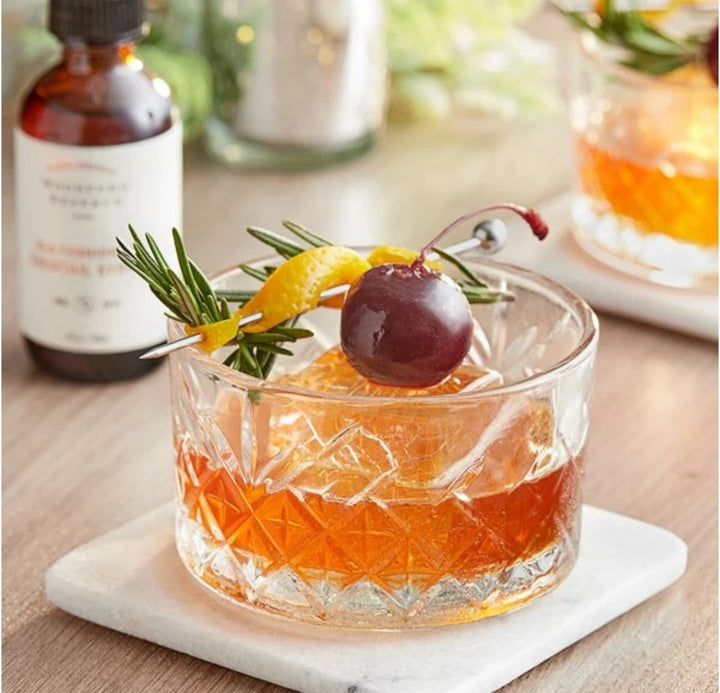 Old Fashioned Cocktail in a glass with garnishes on a marble coaster, with Woodford reserve syrup bottles in the background.