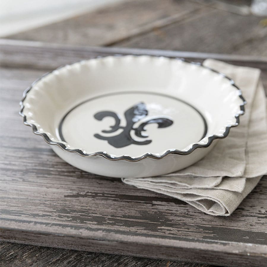 White ceramic pie plate with black rim and black fleur de lis detail on a wooden surface with napkin to the side